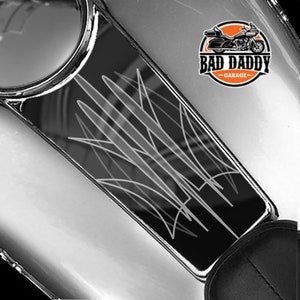 May include: Black motorcycle gas tank cover with a silver pinstripe design. The cover has a logo that says "Bad Daddy Garage".