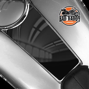 May include: Black motorcycle gas tank cover with a reflective surface and a "Bad Daddy Garage" logo featuring a motorcycle in orange and black.