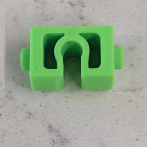 May include: A bright green, 3D-printed plastic component with a U-shaped opening in the center and two small cylindrical protrusions on either side. The object is a solid, geometric shape.