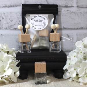 Könnte beinhalten: Drei gläserne Autolufterfrischer mit Holzdeckeln und weißen Holzstäbchen. Die Lufterfrischer sind mit "My Moo Creations Fresh Linen" beschriftet.