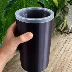 May include: A cylindrical dark purple container with a smooth, matte finish and a translucent white plastic rim at the top. The vessel features a simple, straight-sided design with a wide opening, showcasing a clean and functional aesthetic.
