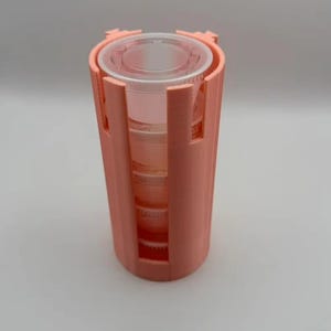 May include: A peach-colored cylindrical holder with vertical cutouts containing a stack of clear, circular plastic containers. The holder features a rigid, textured exterior surface and an open top design that reveals the stacked contents inside.