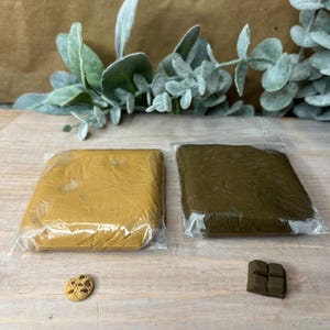 May include: Two blocks of modelling clay, one brown and one yellow, are wrapped in plastic and placed on a wooden surface. A small chocolate bar and a chocolate chip cookie are also on the surface.