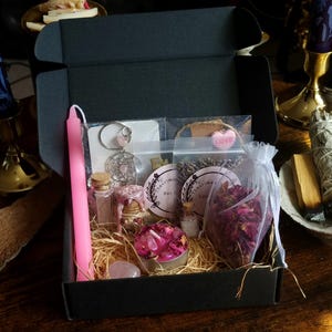 Pode incluir: Uma caixa preta cheia de itens espirituais. Inclui uma vela rosa, pequenos frascos de vidro, um apanhador de sonhos, flores secas, um cristal de quartzo rosa e uma pequena vela com pétalas de rosa. A caixa é disposta sobre palha.