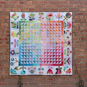 Puede incluir: Una colcha colorida con un patrón geométrico de triángulos en tonos de rosa, naranja, amarillo, verde y azul. La colcha está rodeada por un borde de flores caprichosas y otros diseños.