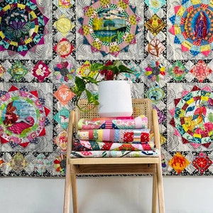 Puede incluir: Una colcha colorida con una variedad de patrones y colores, incluyendo diseños florales, geométricos y abstractos. La colcha está colgada en una pared y está enmarcada por una pared blanca y una alfombra blanca. Una silla de madera con una pila de cuadrados de tela coloridos en el asiento está frente a la colcha.