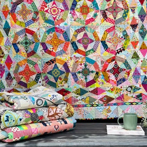 Op de afbeelding: Een stapel kleurrijke vierkanten van stof met geometrische patronen, gevouwen en gerangschikt op een grijs houten oppervlak. Een groot quilt met een vergelijkbaar patroon hangt op de achtergrond.