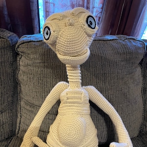 Peut inclure: Un jouet en peluche crocheté du personnage extraterrestre E.T. l'extraterrestre. Le jouet est beige et a de grands yeux noirs avec des pupilles bleues. Le jouet est assis sur un canapé gris.