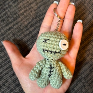 Puede incluir: Llavero de muñeca vudú de crochet verde con un ojo de botón blanco y costuras negras. La muñeca tiene una boca cosida y sostiene un llavero plateado.