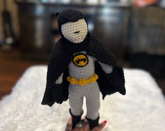 Op de afbeelding: Een gehaakte Batman-pop met een zwarte cape, een gele riem en een grijs pak. De pop staat op een wit, pluche oppervlak.
