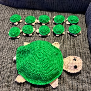 May include: A collection of crocheted green turtles of varying sizes. The largest turtle is lying on its back with its legs and head visible. The smaller turtles are arranged in a semi-circle around the larger turtle.