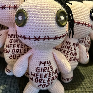 Op de afbeelding: Een set van drie gehaakte poppen met zwart haar en knoopjesogen. Elke pop draagt een roze jurk met rode borduurwerk waarop "My Girls Weekend New Orleans 2023" staat.