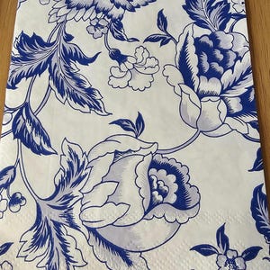 May include: White paper napkin with a repeating blue floral pattern. The design features large flowers, leaves, and smaller floral accents. The pattern is reminiscent of classic blue and white porcelain.