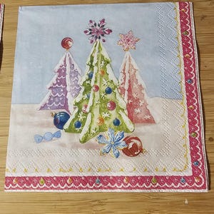 May include: A decorative Christmas-themed paper napkin. The napkin features three decorated Christmas trees in the center, with ornaments and snowflakes. The border is a pink and white scalloped design.
