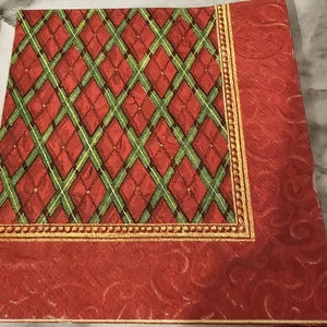 May include: Red and green plaid patterned paper napkin with a gold border. The napkin has a red background with a repeating pattern of green and red diamonds.