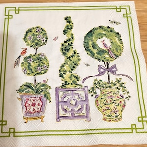 May include: White paper napkin with a green border and a decorative garden scene. The scene features three topiary trees in ornate purple and yellow pots, with birds and butterflies. The napkin has a textured surface.