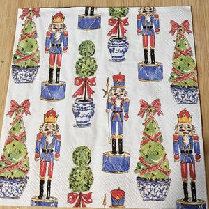 May include: White paper napkin with a repeating pattern of nutcrackers, topiary trees, and decorative pots. The nutcrackers are in blue and red uniforms, and the trees are green with red bows. The pots are blue and white.