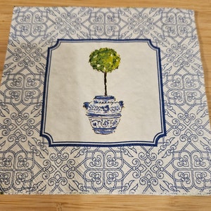May include: A white napkin with a blue and white patterned border. The center of the napkin features a blue and white ceramic pot with a green topiary tree.