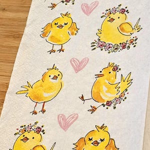 May include: A white kitchen towel with a repeating pattern of yellow chicks wearing floral crowns and pink hearts. The chicks are drawn in a whimsical style with simple lines and bright colors.