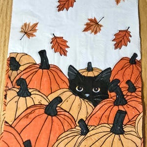 May include: A decorative fall-themed napkin featuring a black cat peeking out from behind a collection of orange and yellow pumpkins. Brown autumn leaves are scattered across the white background. The pumpkins have black stems.