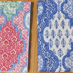 May include: Two square napkins featuring a repeating ogee pattern. The left napkin displays pink, light blue, and green floral motifs. The right napkin features a monochromatic blue and white design with intricate botanical and geometric details.