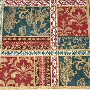 May include: A decorative square napkin with a patchwork design. The design features floral and paisley patterns in teal, burgundy, and gold. Borders and vertical stripes are in coordinating colors. The napkin is likely made of paper.