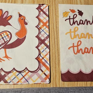 May include: Two Thanksgiving-themed paper napkins. One has a cartoon turkey in orange, brown, and cream colours. The other has the word "thankful" in a script font, with autumn leaves.