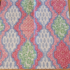Op de afbeelding: Een vierkant stoffen servet met een herhalend vierpaspatroon in tinten roze, groen en blauw. Het ontwerp bevat ingewikkelde bloemmotieven en geometrische ruitvormen gedrukt op een witte achtergrond met een blauwe omlijnde rand.