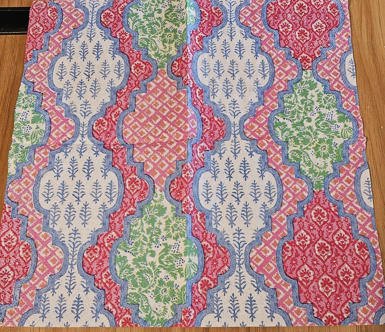 Op de afbeelding: Een vierkant stoffen servet met een herhalend vierpaspatroon in tinten roze, groen en blauw. Het ontwerp bevat ingewikkelde bloemmotieven en geometrische ruitvormen gedrukt op een witte achtergrond met een blauwe omlijnde rand.
