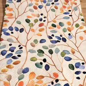 May include: A white paper napkin with a floral pattern. The pattern features branches with small leaves in shades of blue, green, orange, and yellow.