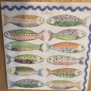 May include: A white paper napkin with a blue wavy border and 12 colorful fish illustrations. The fish are all different colors and patterns, including red, green, blue, yellow, orange, and purple. The napkin is decorated with the text "Rockstar Grill" in blue.