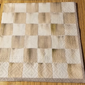 May include: A square paper napkin with a checkered pattern in shades of beige and white. The border features a repeating diamond design. The napkin is likely for dining or decorative use.