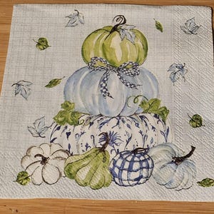 May include: A stack of four pumpkins in various shades of blue and green with a white and blue patterned pumpkin at the bottom. The pumpkins are decorated with leaves and a plaid ribbon.