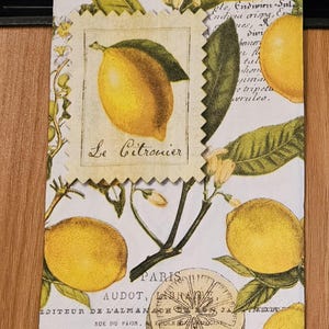 May include: A rectangular paper napkin featuring a botanical lemon print with green leaves and white blossoms. The design includes the text Citrus, Le Citronier, and vintage French typography. A central square patch with a lemon image is layered on top.