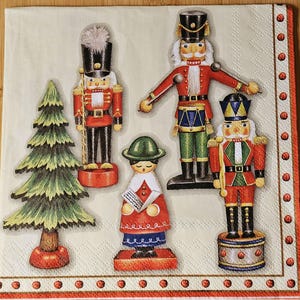 May include: A decorative napkin featuring a festive Christmas theme. The design includes illustrations of a Christmas tree, a singing figure, and several nutcracker soldiers in red, black, and gold uniforms. The napkin's border is adorned with red and white accents.