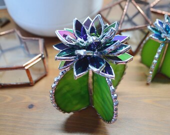 May include: A stained glass cactus with a green body and a purple and green flower. The cactus is made of glass and has a silver trim.