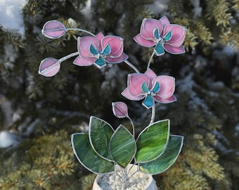 May include: A stained glass orchid plant featuring pink and teal petals with green leaves, set in a white geometric ceramic pot filled with white pebbles. The structure is supported by thin metal stems, creating a decorative floral display.