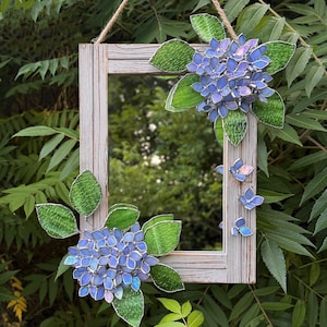 May include: A rectangular mirror with a weathered, gray wooden frame, adorned with blue and purple stained glass hydrangea flowers and green leaves. The mirror is suspended by a natural rope, creating a decorative garden accent.