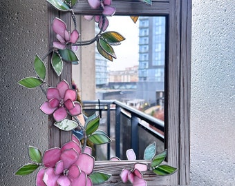 May include: A stained glass mirror with a wooden frame decorated with pink and green flowers. The mirror reflects a city view.