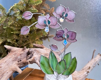 May include: A stained glass orchid plant featuring light pink and purple petals with iridescent blue centers, green glass leaves, and silver metal soldering. The plant is potted in a white geometric ceramic container filled with white stones.