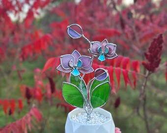 May include: A stained glass orchid plant featuring purple and teal petals with green leaves, held in a white geometric ceramic pot filled with small white stones. The glass pieces are joined by silver metal solder lines, creating a floral sculpture.