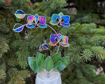 May include: A stained glass orchid plant featuring iridescent blue and purple petals with dark centers, green glass leaves, and a small bud. The plant is set in a white geometric ceramic pot filled with small white stones, creating a decorative piece.
