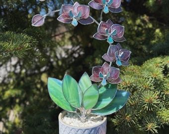 May include: A stained glass orchid plant featuring purple and teal petals with green leaves, set in a textured light blue ceramic pot filled with white pebbles. The plant has a curved stem with multiple blooming flowers and several buds.