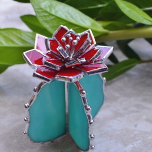 Könnte beinhalten: Ein Kaktus aus Buntglas mit einer roten Blüte oben. Der Kaktus ist aus türkisfarbenem Glas und hat silberne Akzente.