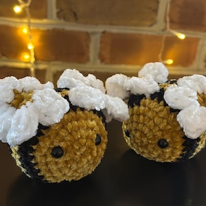 May include: Two crocheted bee plushies with black and yellow bodies and white fuzzy flower accents.