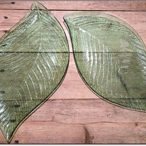 May include: Two leaf-shaped glass plates with a textured, veined design. The plates are a pale green colour and sit on a wooden surface. The plates are decorative and suitable for serving food.