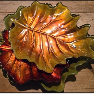 Puede incluir: Una pila de platos decorativos de vidrio con forma de hoja. Los platos son en tonos ámbar, dorado y rojo, con un acabado brillante. Los platos con forma de hoja están dispuestos sobre una superficie de madera, creando una exhibición con temática otoñal.