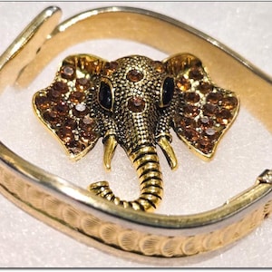 May include: A gold-tone bangle bracelet with a detailed elephant charm. The elephant is adorned with brown rhinestones and has a black enamel eye.