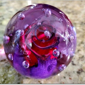 Op de afbeelding: Een heldere glazen presse-papier met een levendig interieurontwerp. De presse-papier heeft een wervelend rood en paars bloemmotief, met luchtbellen die in het glas zweven. De buitenkant is doorschijnend paars.