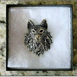 May include: A detailed brooch in the shape of a wolf's head, presented in a black box with white padding. The brooch features black and silver-toned crystals, with gold accents on the nose and eyes. The wolf's head is centered in the box.